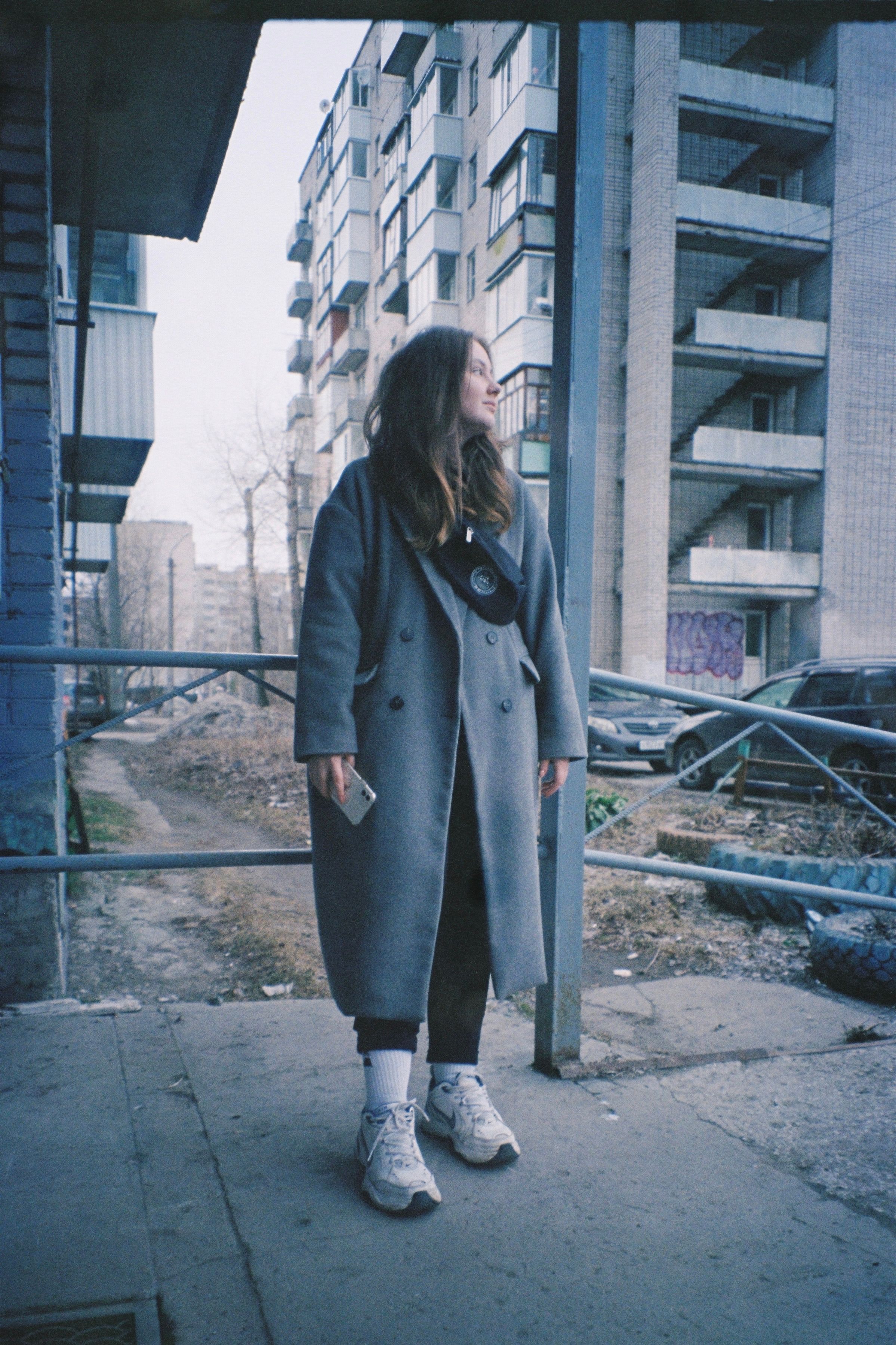 Young woman in a gray coat standing confidently in an urban setting.