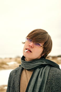 Portrait of a young adult with eyeglasses in winter clothing smoking outdoors.