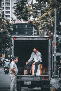 Two Asian men communicate near a parked truck on a city street, surrounded by trees and buildings.
