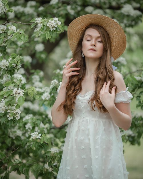 Free A serene portrait of a woman in a white dress and straw hat, surrounded by blossoming trees in spring. Stock Photo