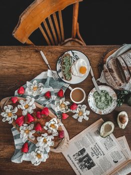 Cozy breakfast setup with avocado toast, fried egg, strawberries, and coffee on a wooden table.