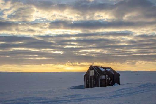 A serene winter sunrise over a snow-covered landscape featuring a solitary wooden hut under dramatic clouded skies.