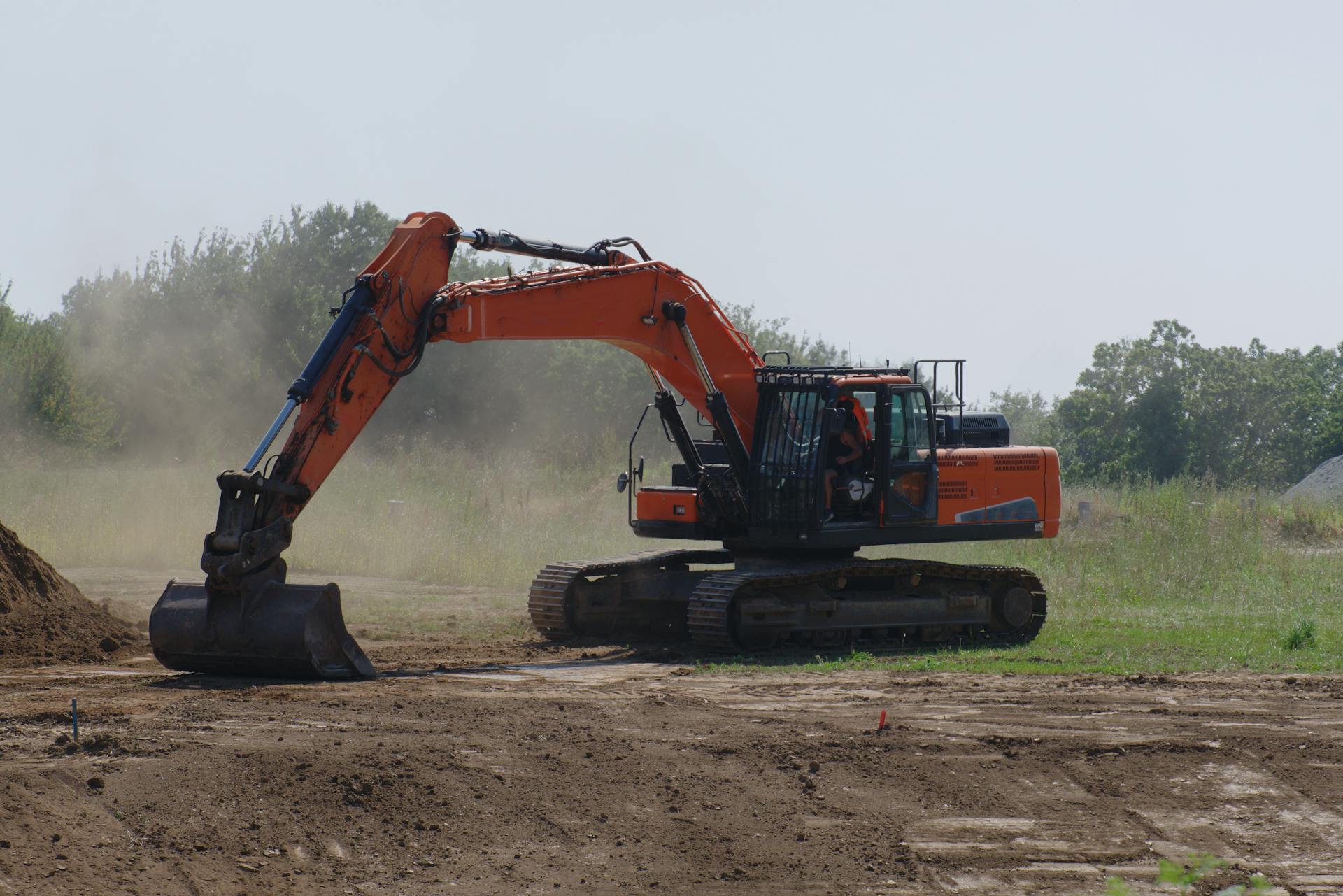 An excavator levels dirt at an outdoor construction site with dust and trees visible.