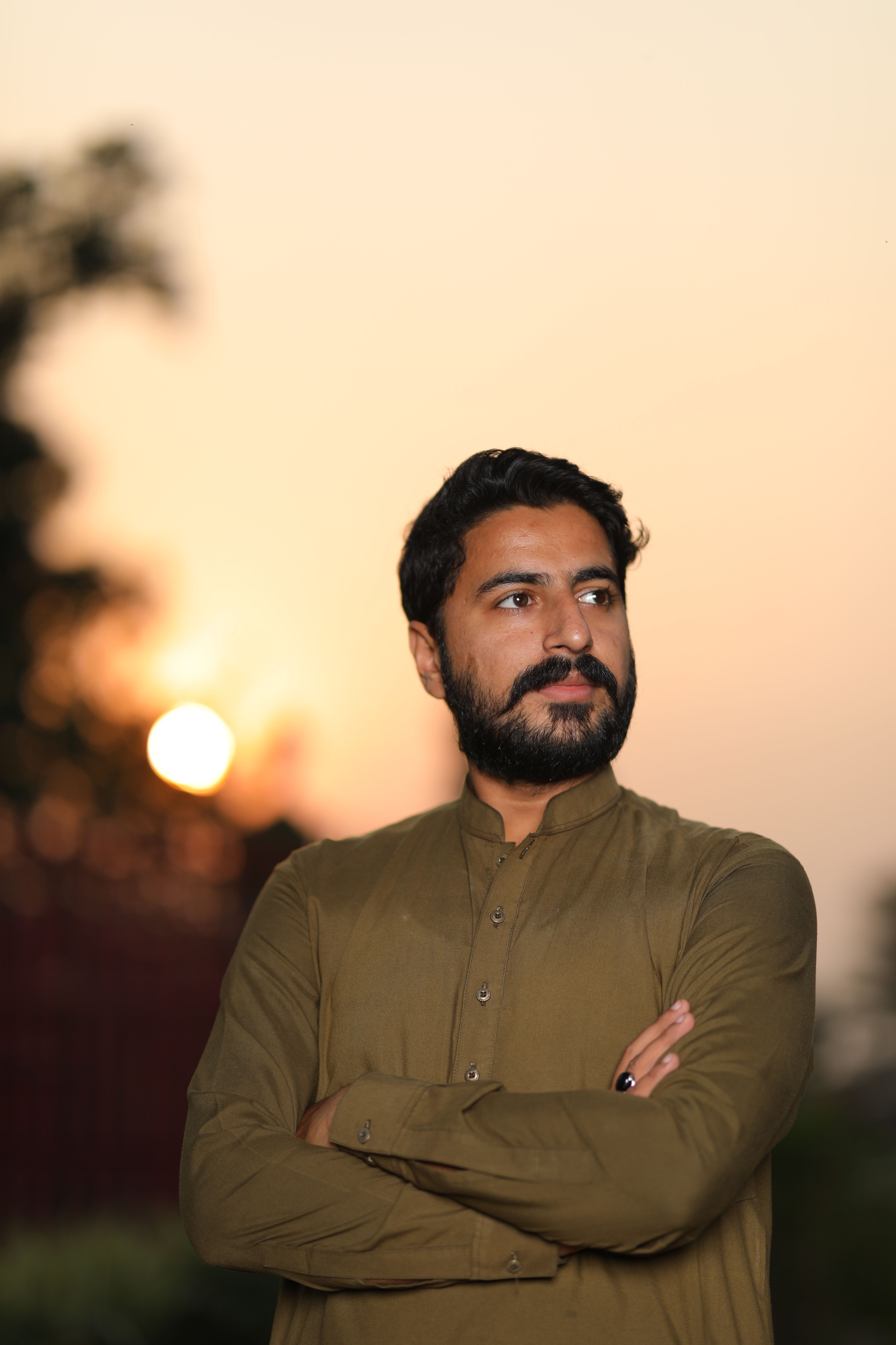 Close-up portrait of a bearded man in brown long sleeves with folded arms at sunset.