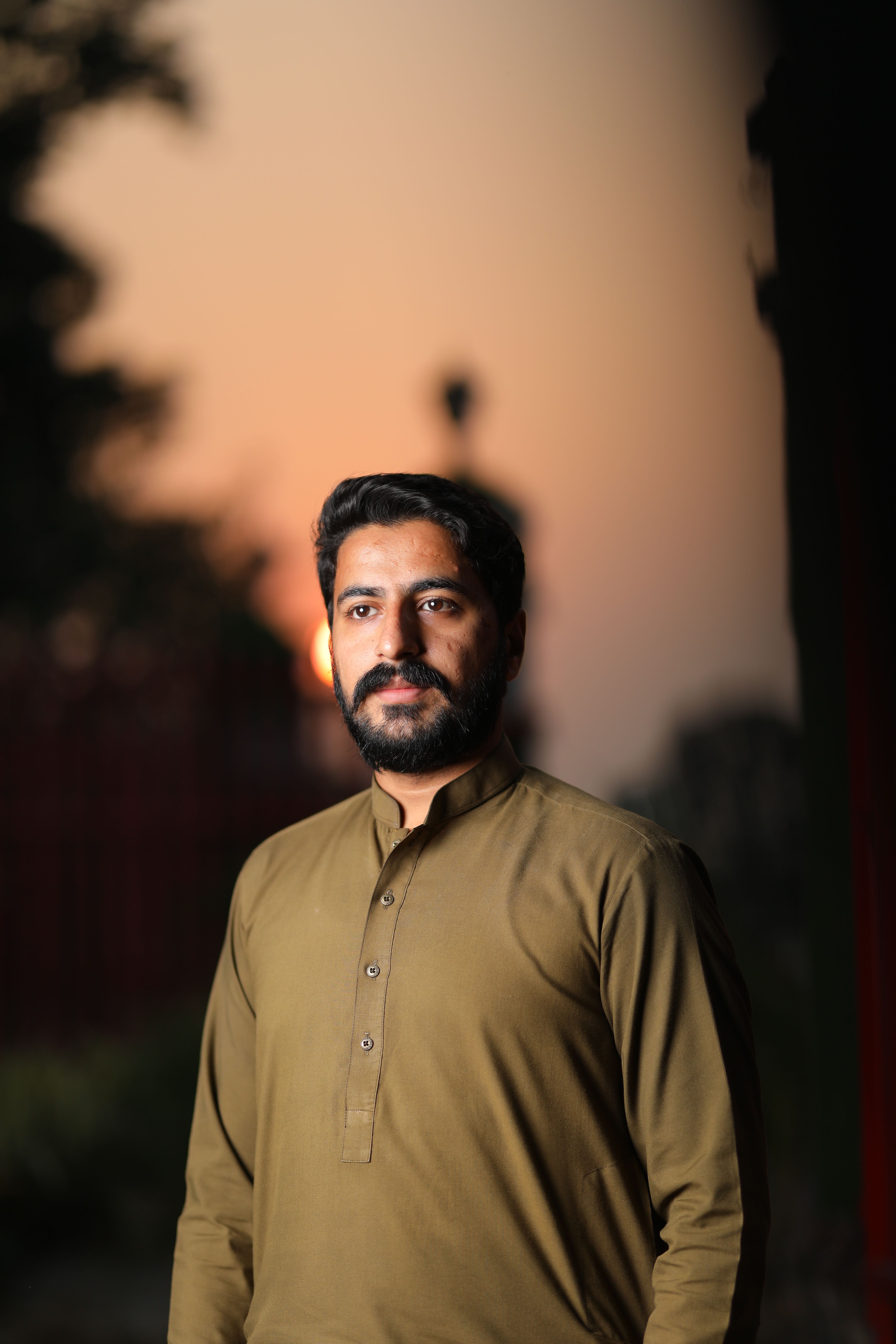 A bearded man in a traditional shirt standing outdoors at sunset, with a soft focus background.