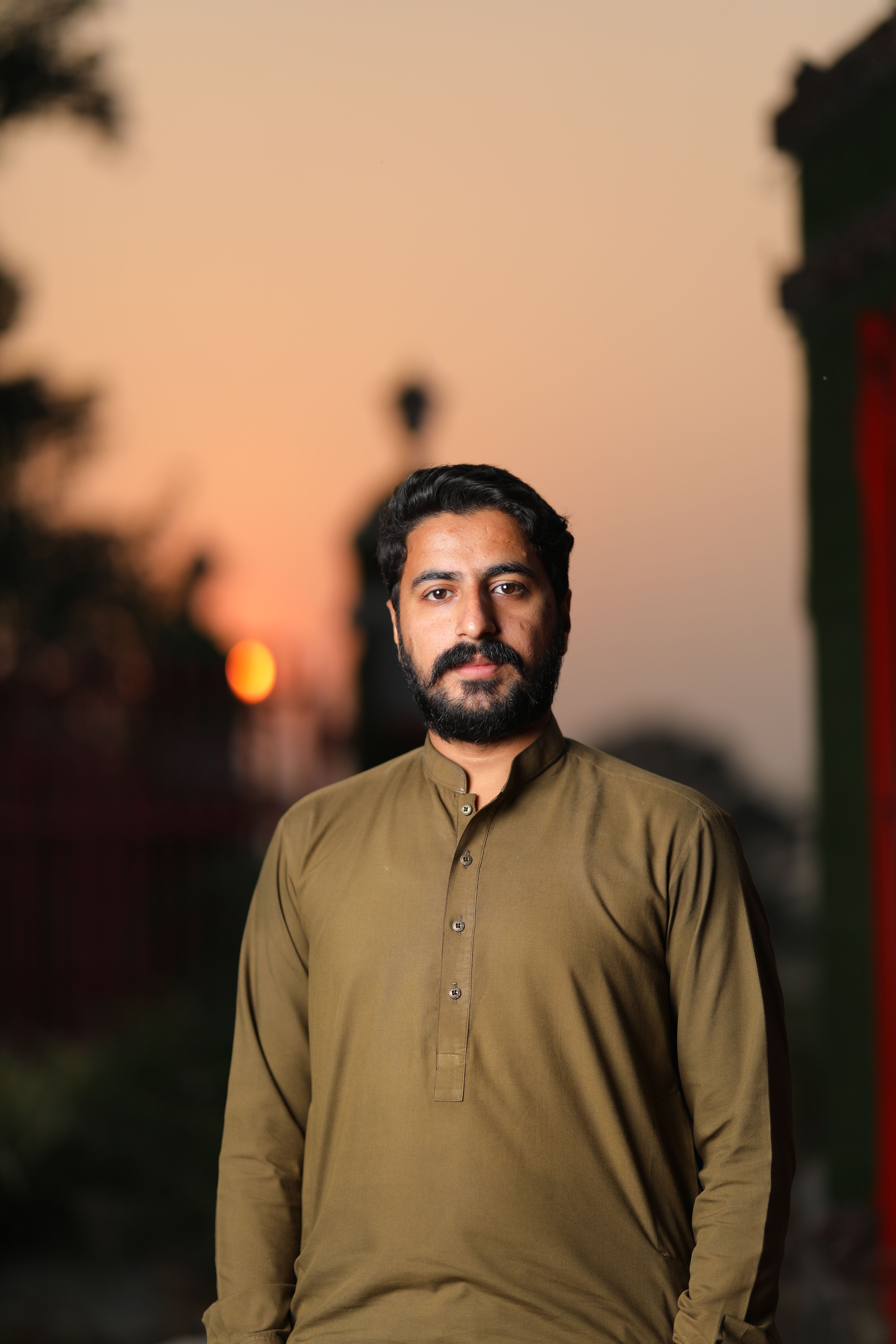 A bearded man in green long sleeves stands outdoors with a sunset backdrop.
