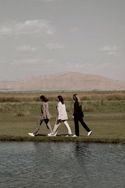 Three women in fashionable attire walking by a river with mountains in the background, set in a serene landscape.
