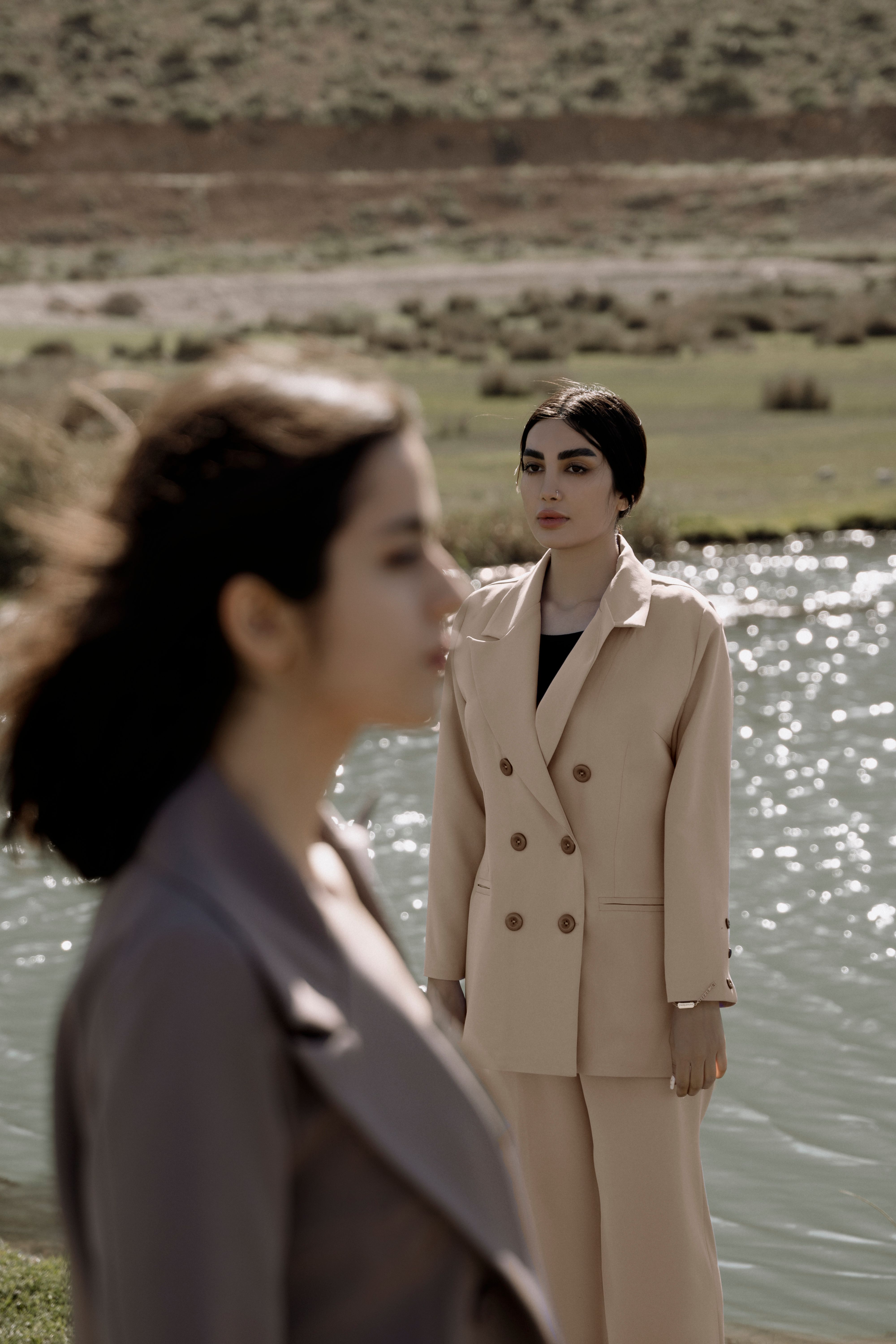 Two women in stylish suits stand by a tranquil lake, exuding elegance and poise.