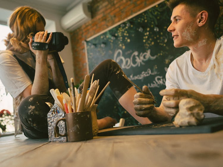 Woman On Table Filming Guy Working In Clay