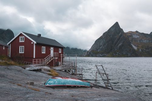Free Quaint red cabin by Norwegian fjord with dramatic mountain backdrop under cloudy skies. Stock Photo