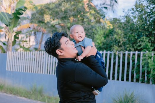 A happy father lifts his smiling baby in a sunny outdoor setting, capturing a joyful family moment.