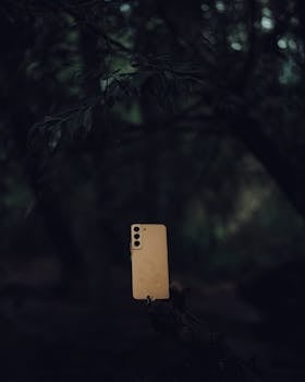 A smartphone covered in raindrops set against a dark, forest background.
