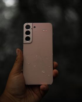 A hand holds a water-dropped smartphone against a blurred background.
