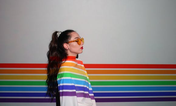 A woman wearing sunglasses stands by a rainbow-striped wall, symbolizing pride and diversity.