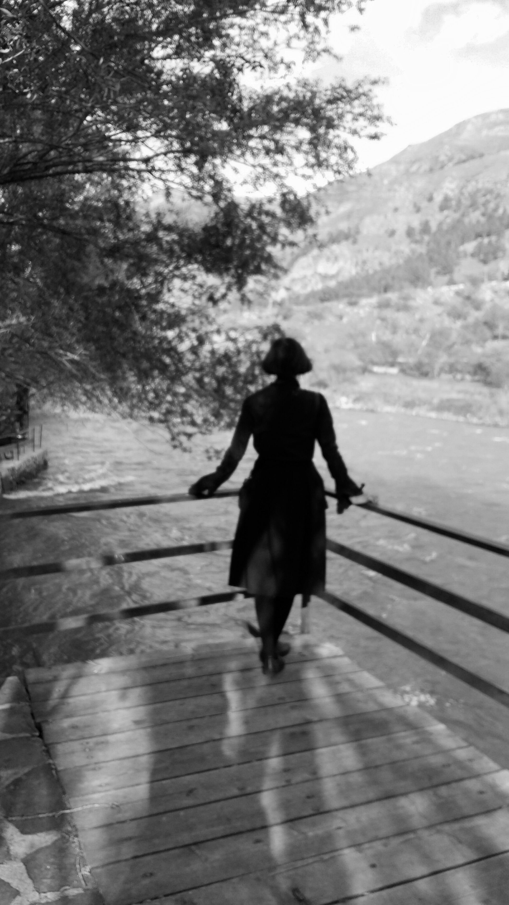 Silhouette of a woman in a black dress standing on a wooden terrace, gazing at a mountain river.