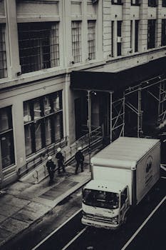 Monochrome image of a truck on a New York City street with pedestrians nearby.
