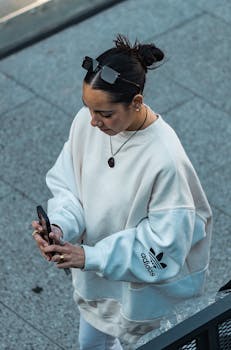Young woman using smartphone outdoors, wearing a white sweater and sunglasses.