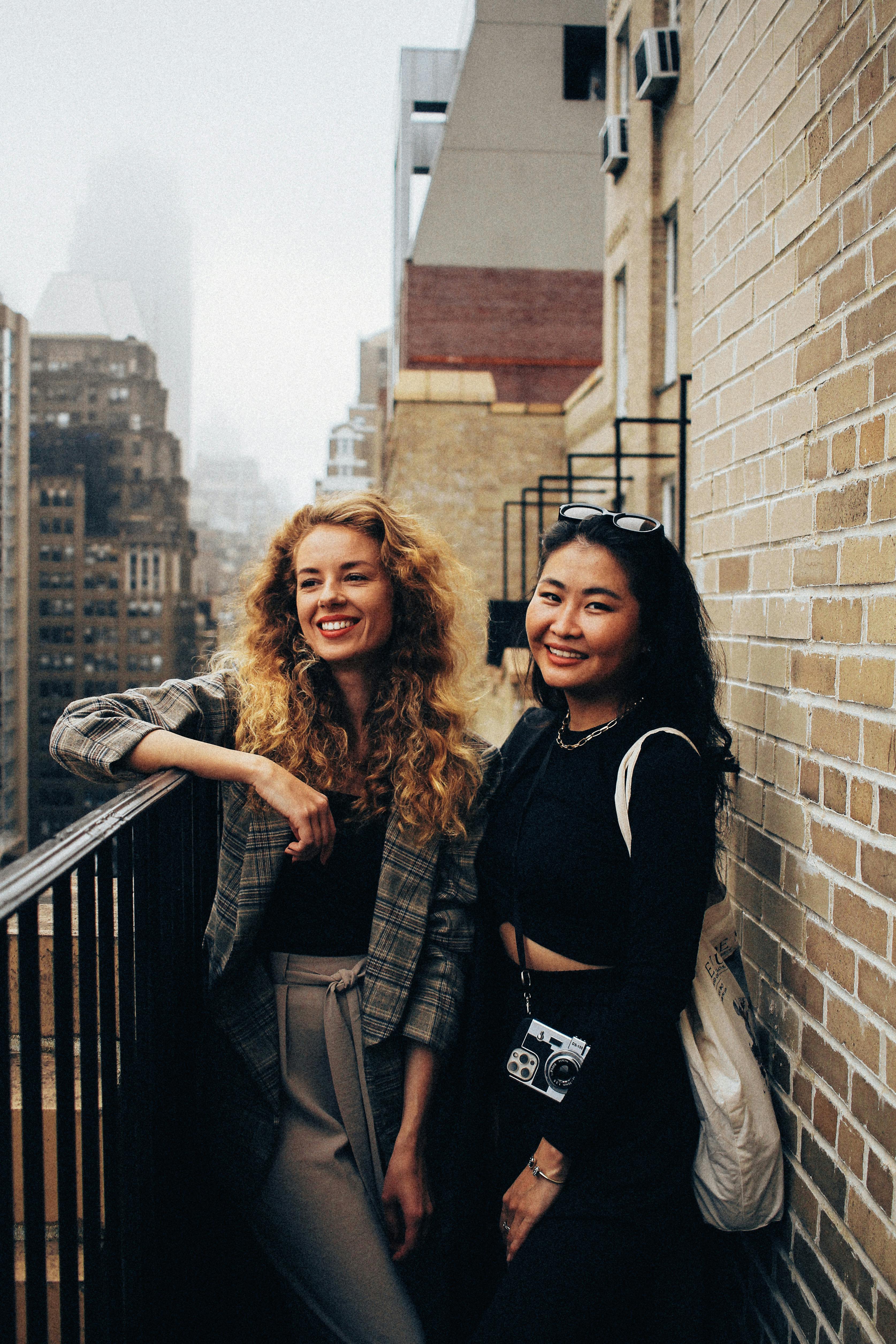 Two women smiling on a New York balcony with city skyline in the background.