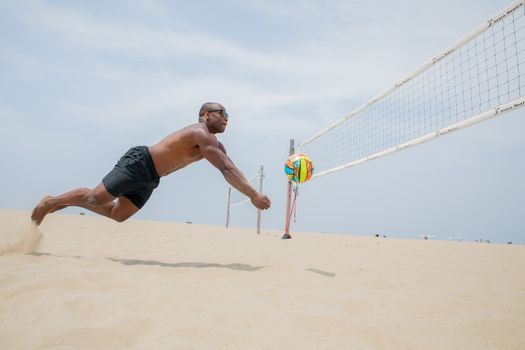 A muscular man dives for the volleyball during a game on a sunny beach, showing athleticism and energy.
