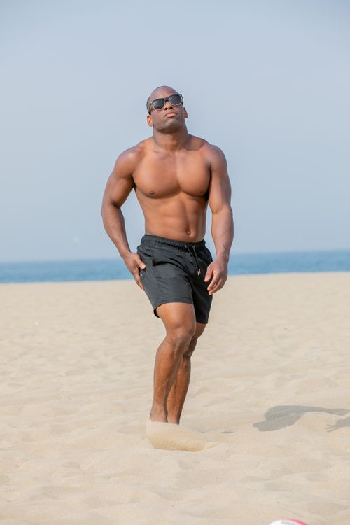 Free Shirtless man with sunglasses walking on a sandy beach under clear skies. Stock Photo