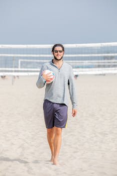 A young man with sunglasses holds a volleyball on a sandy beach, near a net, under clear skies.
