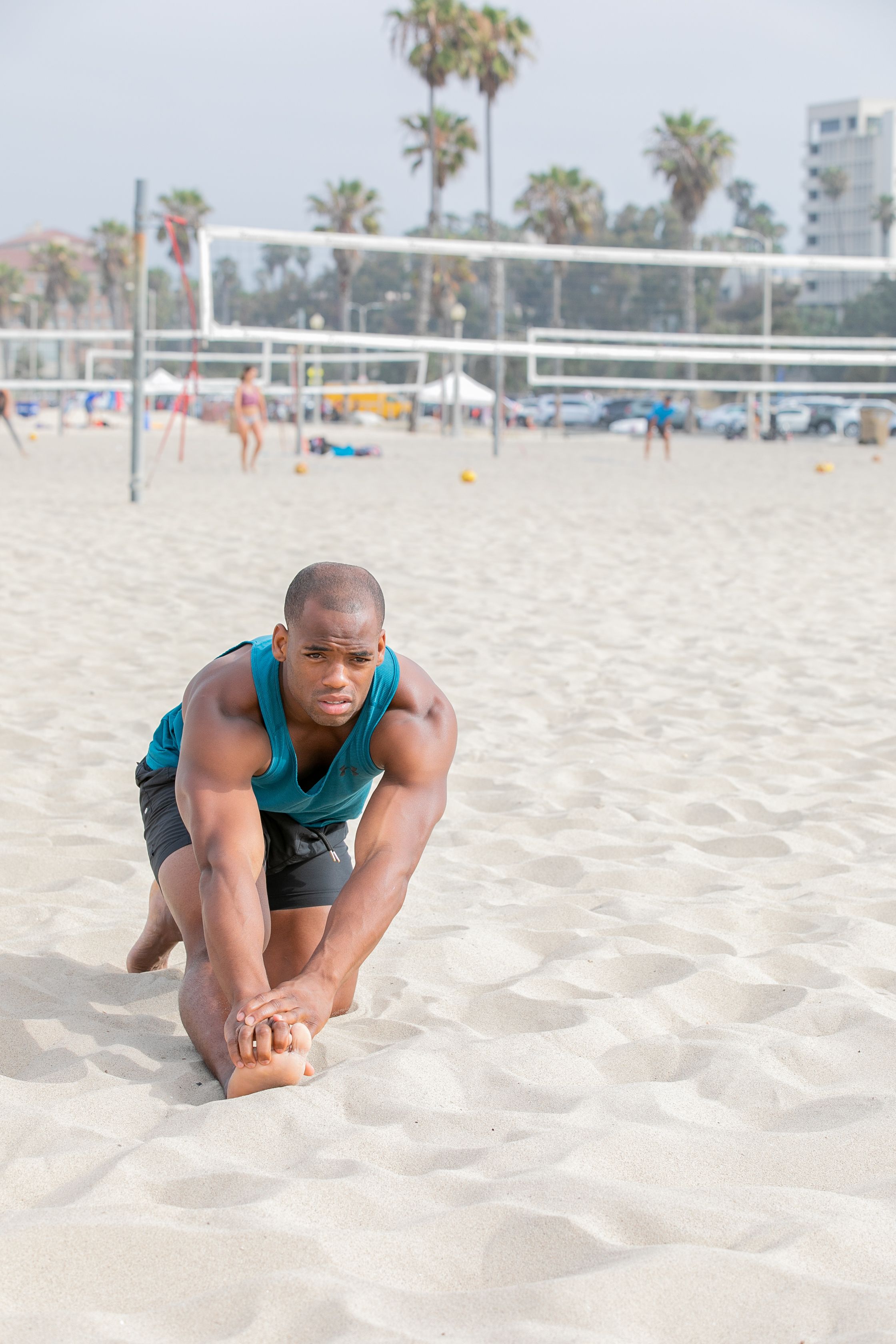 Muscular man performing stretching exercises on a beach volleyball court in daylight.