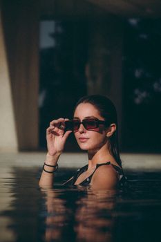Elegant woman in a swimming pool wearing sunglasses, posing confidently.
