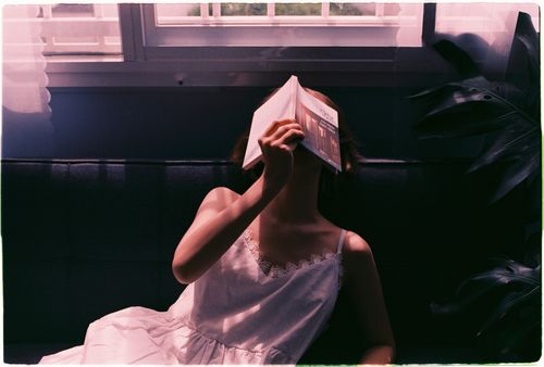 Free A woman in a white dress sits on a couch, covering her face with a book indoors with sunlight. Stock Photo