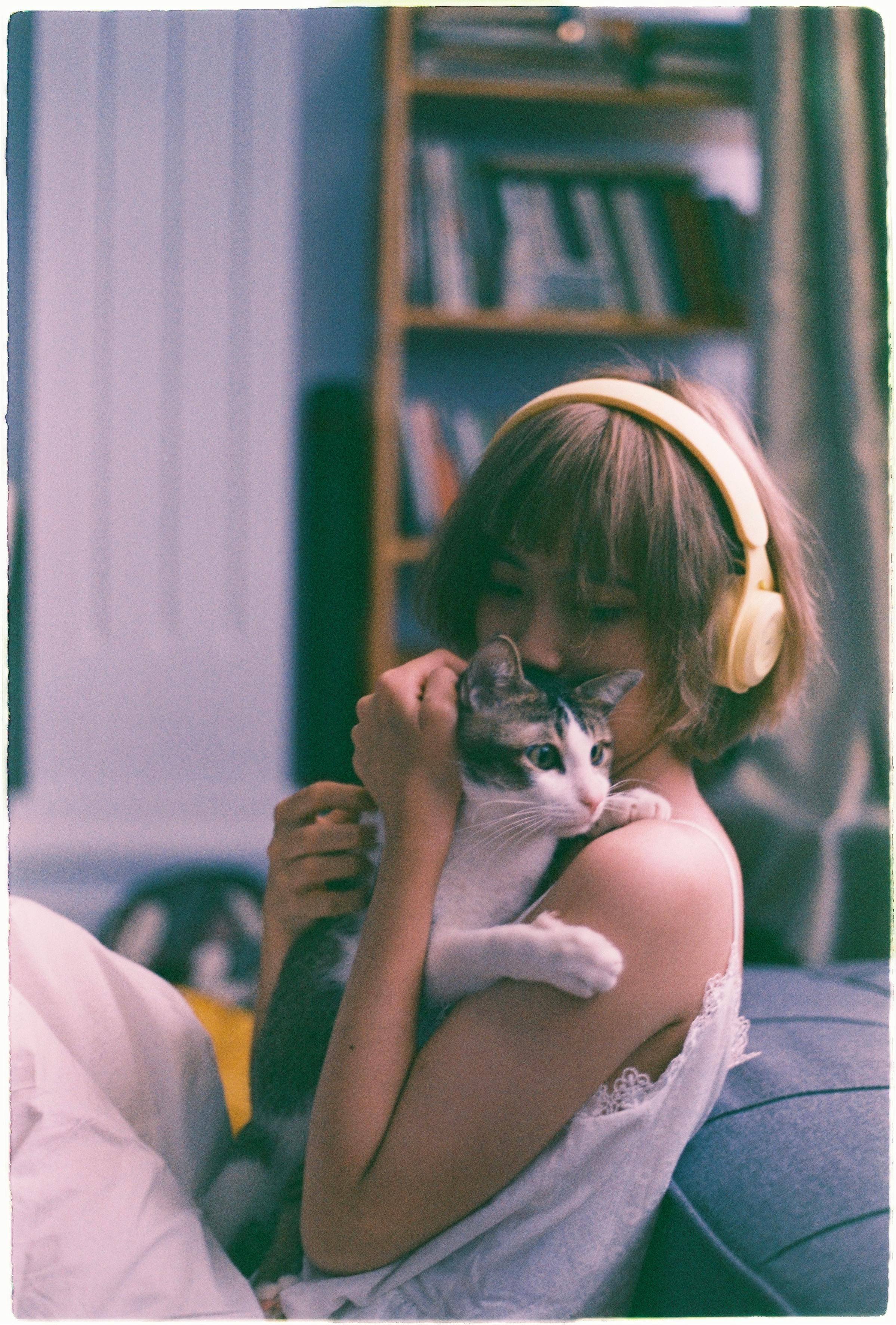 A young woman with headphones holds a cat, enjoying a peaceful moment indoors.