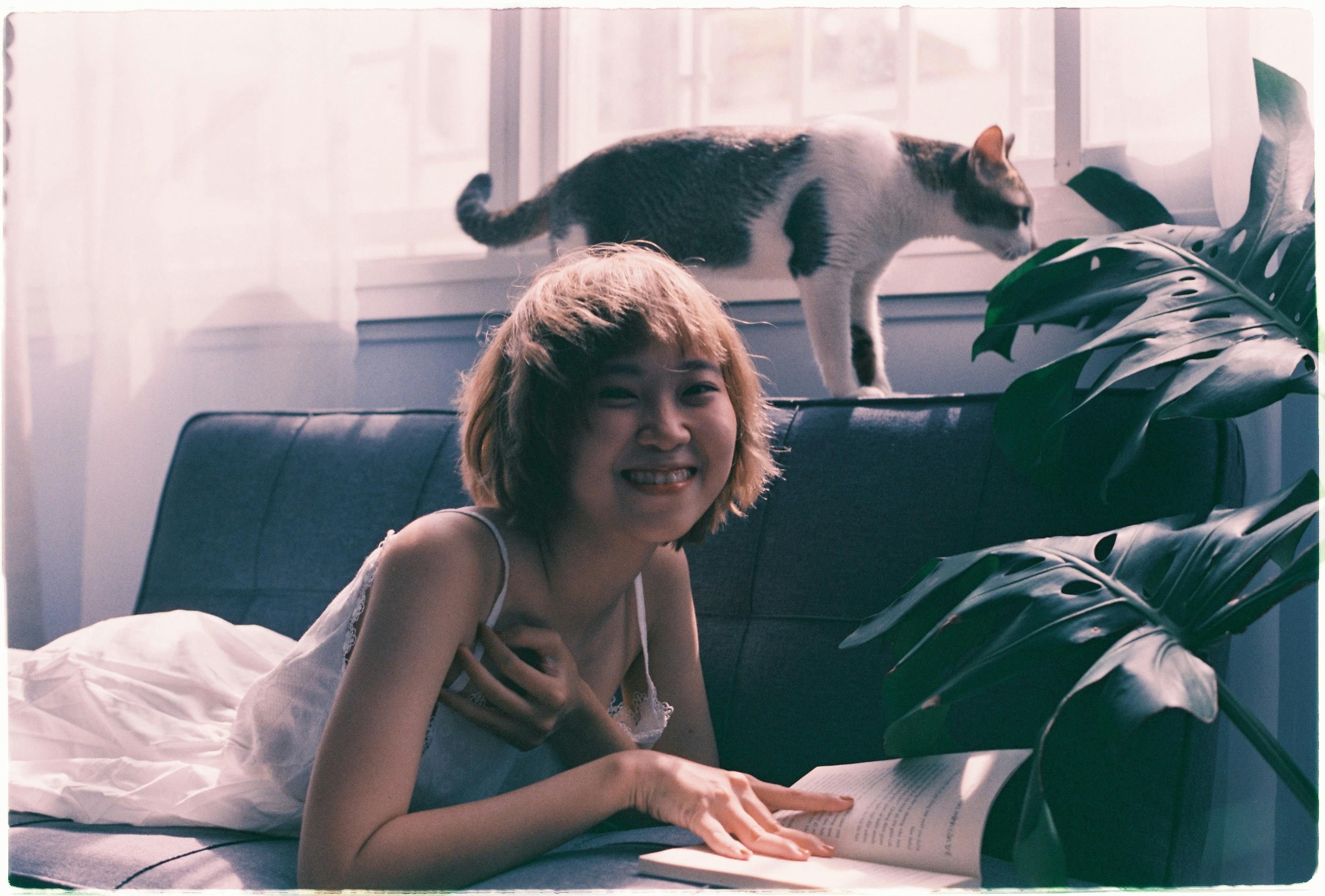 Smiling Asian woman in white dress reading a book on a sofa with her cat nearby.