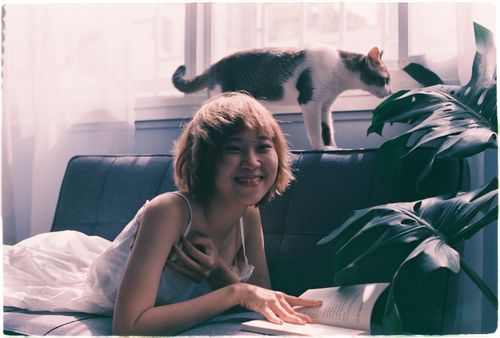 Free Smiling Asian woman in white dress reading a book on a sofa with her cat nearby. Stock Photo