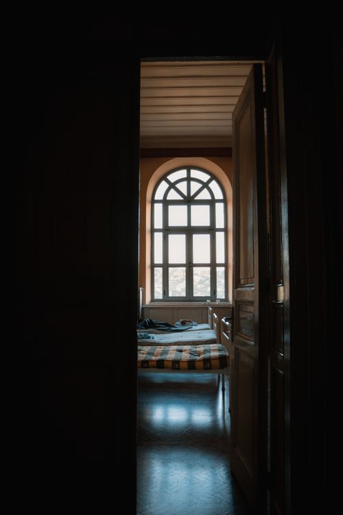 Free A dimly lit bedroom seen through an open wooden door with a large window. Stock Photo