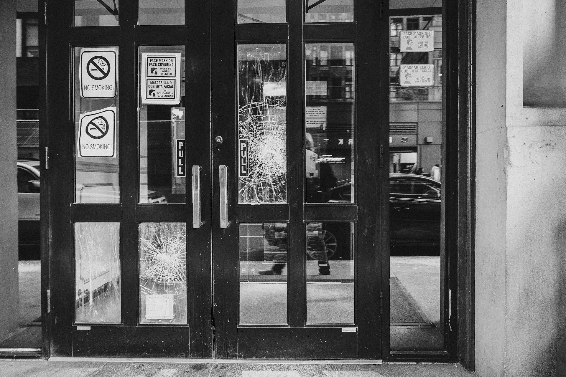 A grayscale image of a cracked glass door with signage and reflections, creating a moody urban scene.