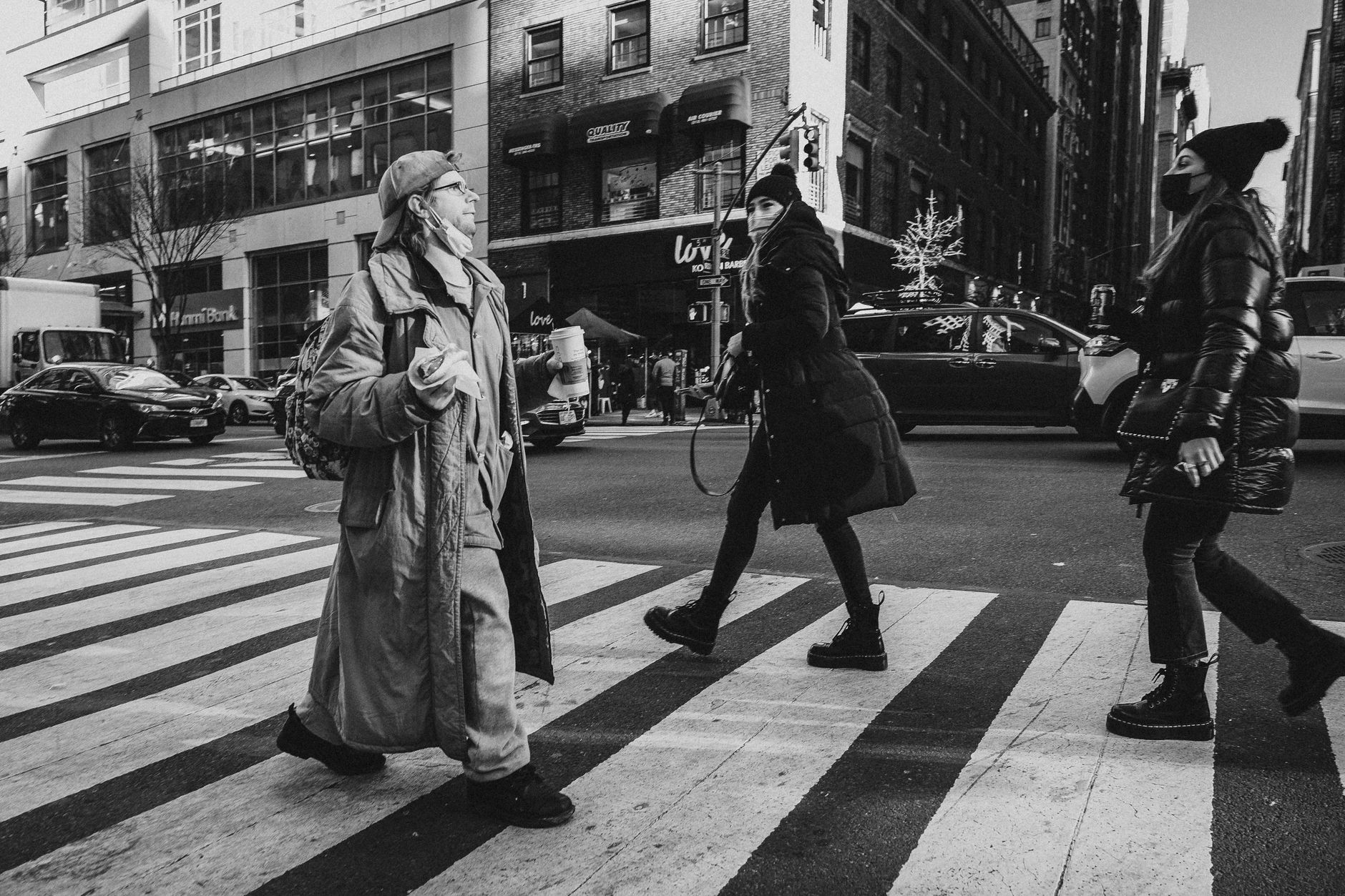 Black and white photo of people crossing an urban street, wearing winter clothing and facemasks.
