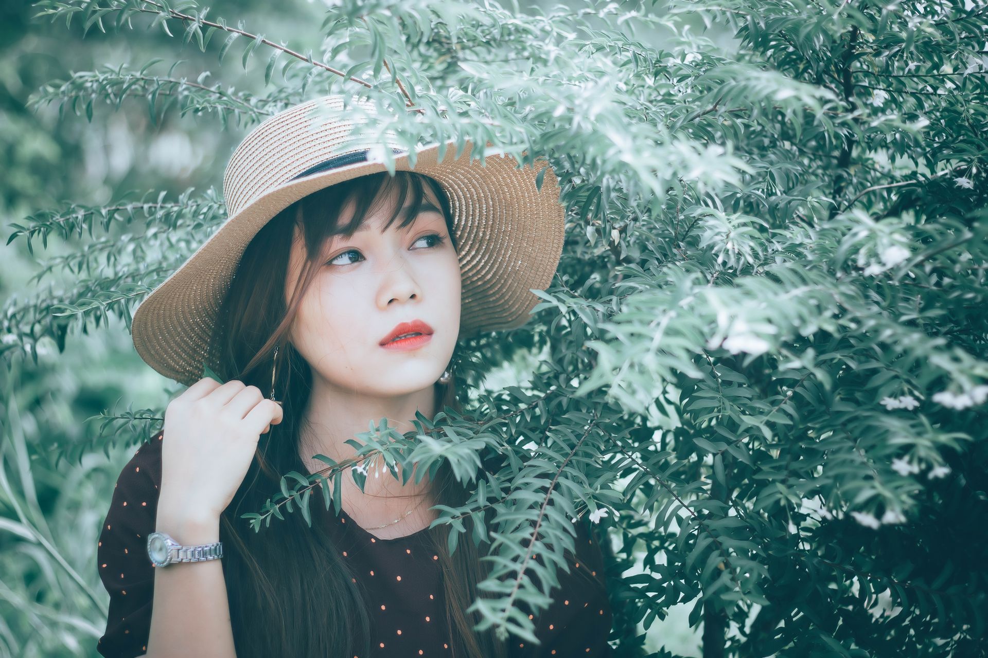 A beautiful Asian woman poses gracefully among lush greenery, wearing a stylish straw hat.