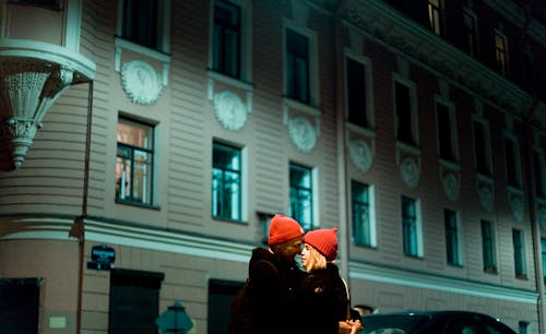 戴著紅色針織帽的男人和女人在夜間親吻在灰色的混凝土建築前