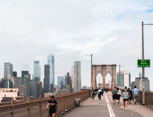 Free People walking on Brooklyn Bridge with Manhattan skyscrapers in the background under cloudy skies. Stock Photo