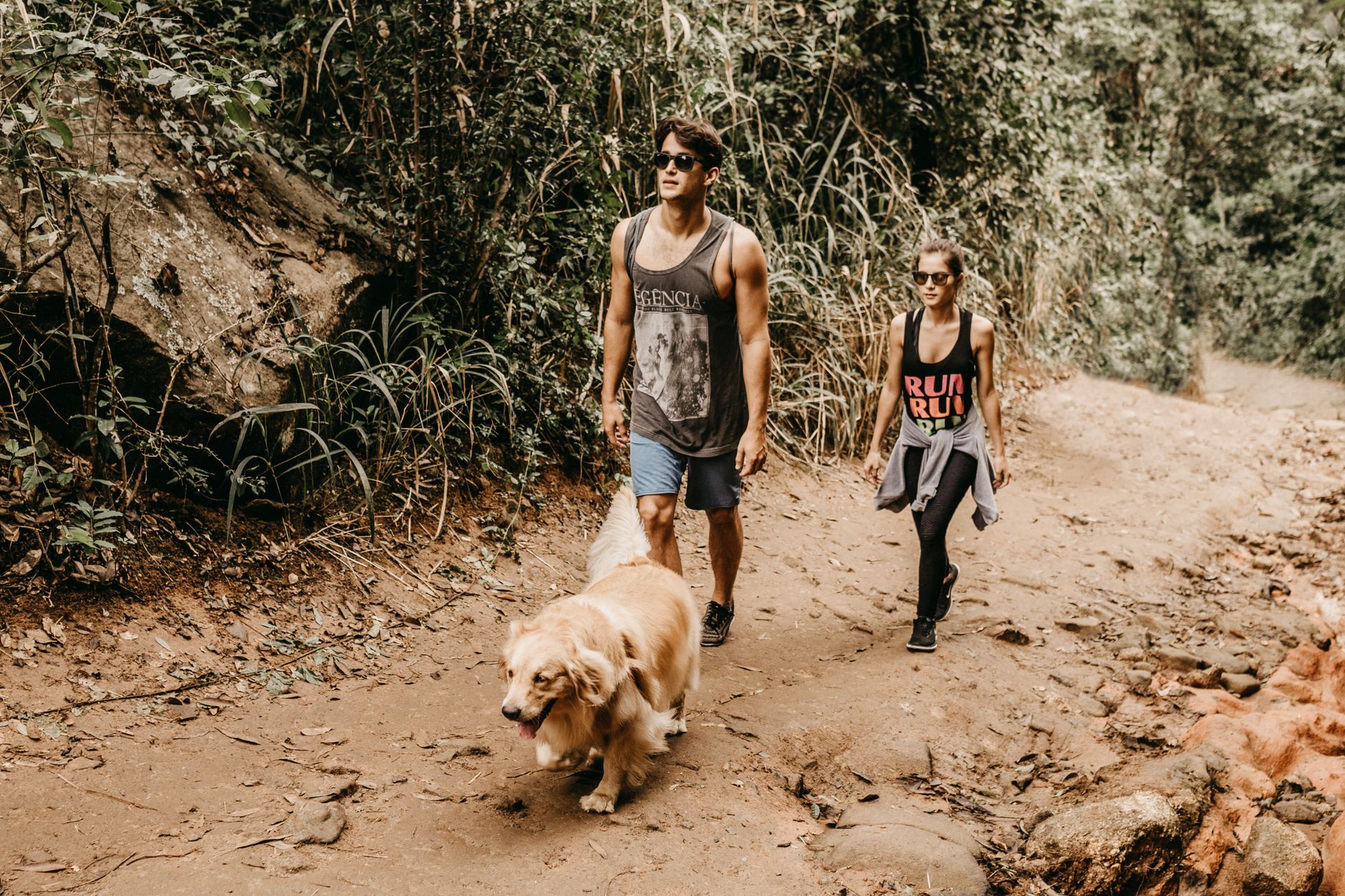 A couple walks their golden retriever along a scenic dirt trail in a lush forest setting.