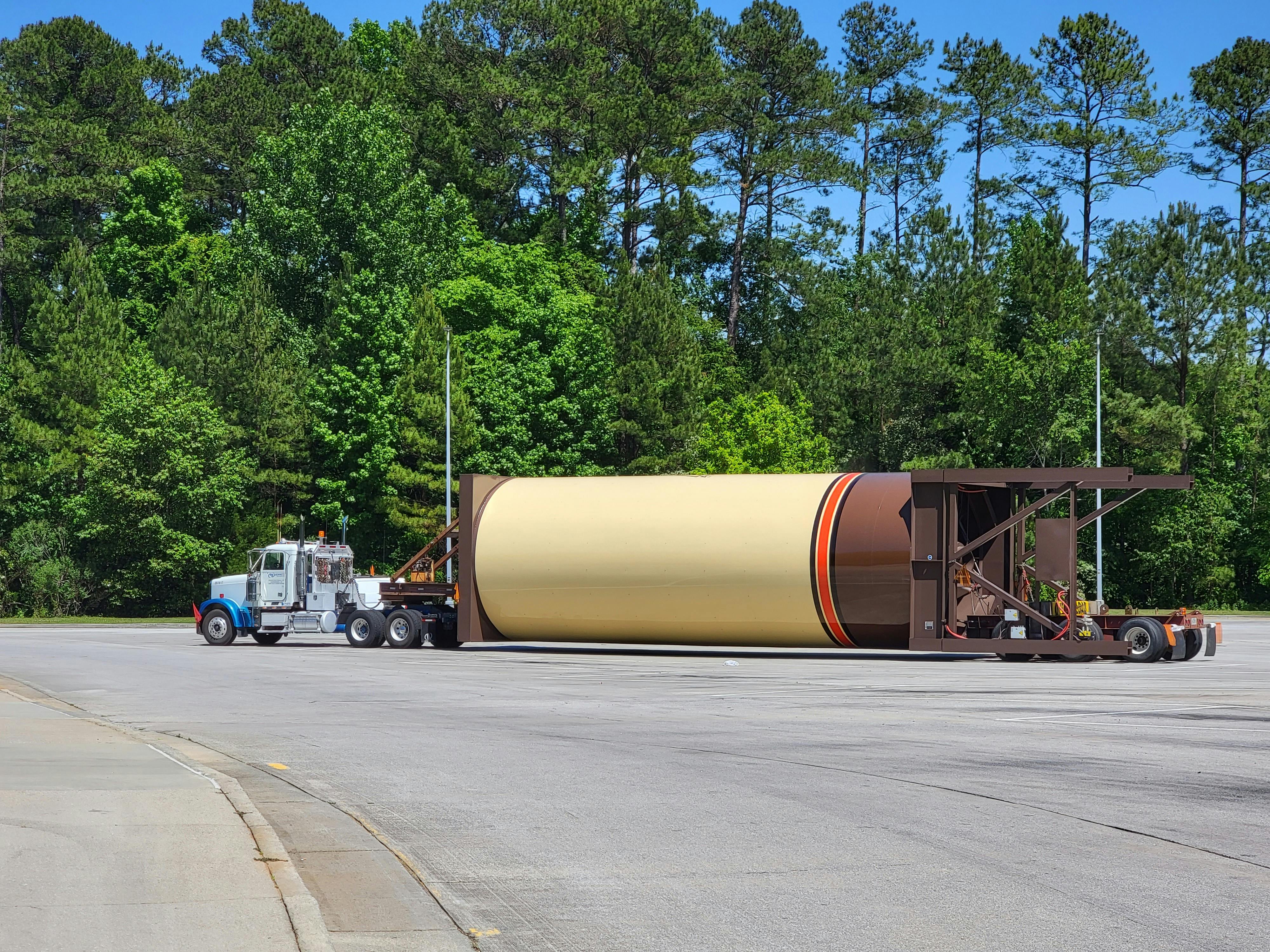 A heavy-duty truck transports large cylindrical equipment on an outdoor road beside lush forests.