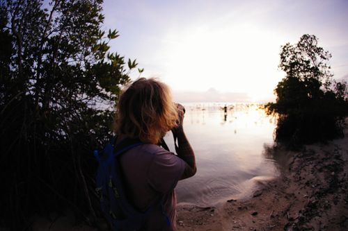 Free A photographer with backpack captures a scenic sunset by a tranquil lake shoreline. Stock Photo