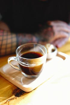 A hot cup of coffee in a glass mug on a wooden tray with a tattooed arm in the background.