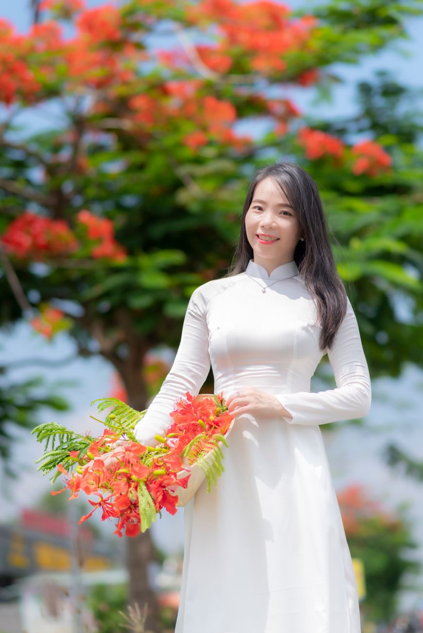 Asian woman in traditional Ao Dai holding flame tree flowers outdoors, showcasing elegance.