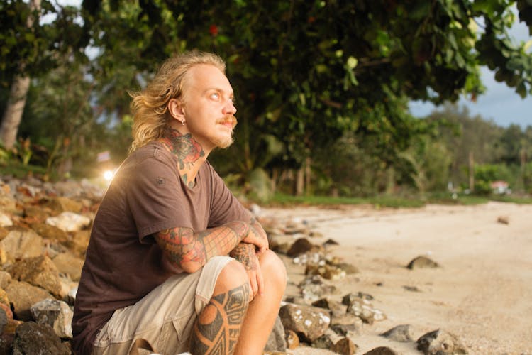 Man With Tattoos Sitting On The Rocks On The Shore