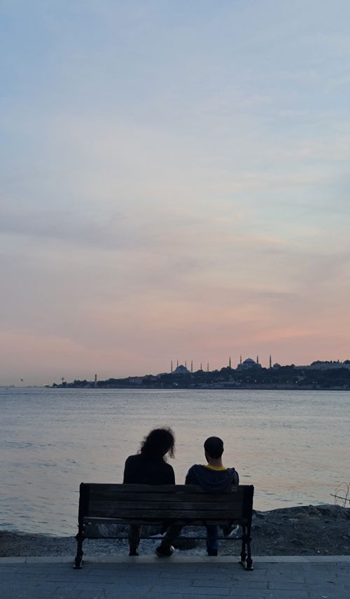 Free Two individuals sitting on a bench enjoying a peaceful sunset by the water, silhouetted cityscape in view. Stock Photo