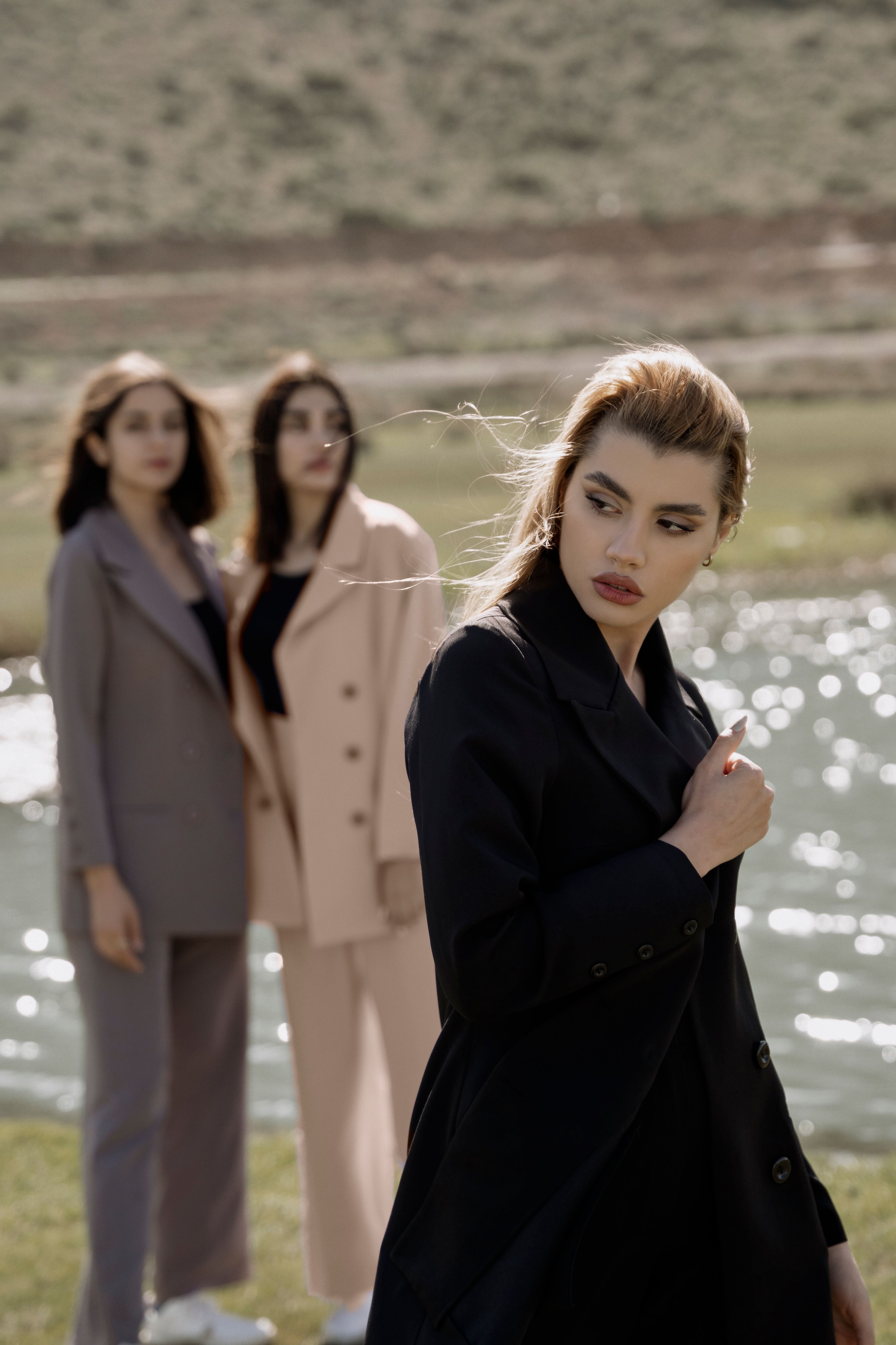Three women in fashionable coats pose by a sunlit river, showcasing elegance and style.