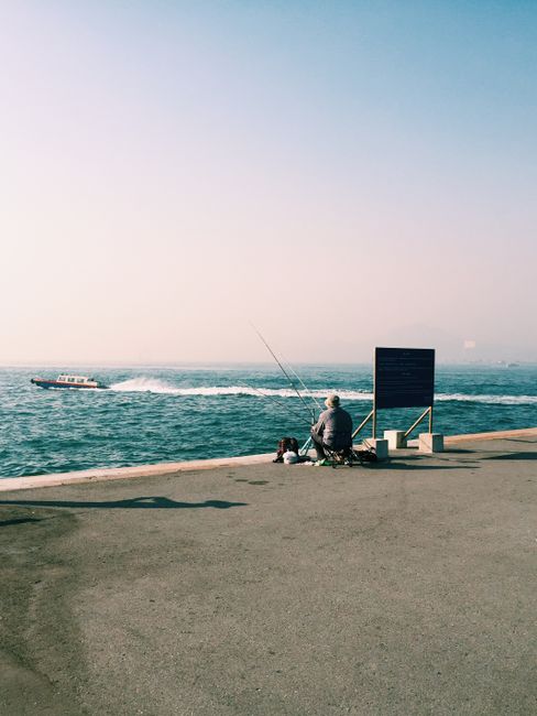A lone fisherman enjoys a peaceful day by the sea in Kowloon, Hong Kong, with a boat passing by.