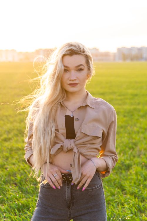 Free Portrait of a blonde woman posing confidently in a sunlit field, with long hair and casual attire. Stock Photo