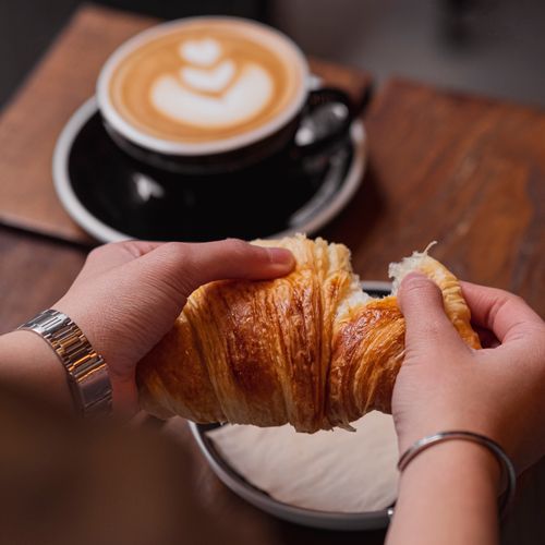 Free Hands breaking a croissant with a latte in the background, showcasing a cozy coffee shop vibe. Stock Photo