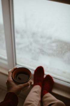 A person relaxes by a window, sipping coffee, wearing red socks, while enjoying a snowy view.