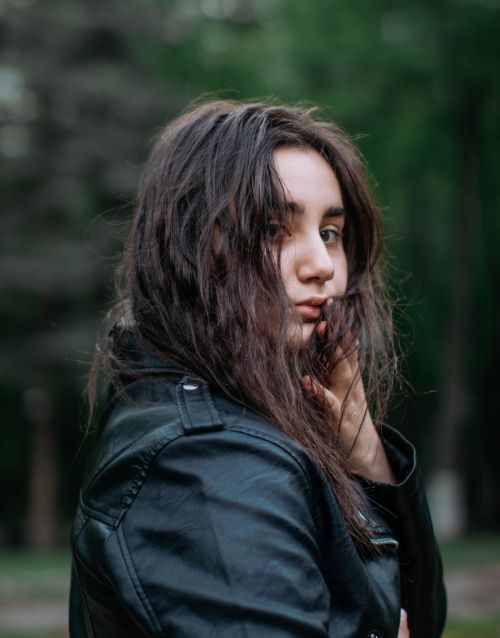 Free Portrait of a young woman with long hair in leather jacket, set against a lush green outdoor background. Stock Photo
